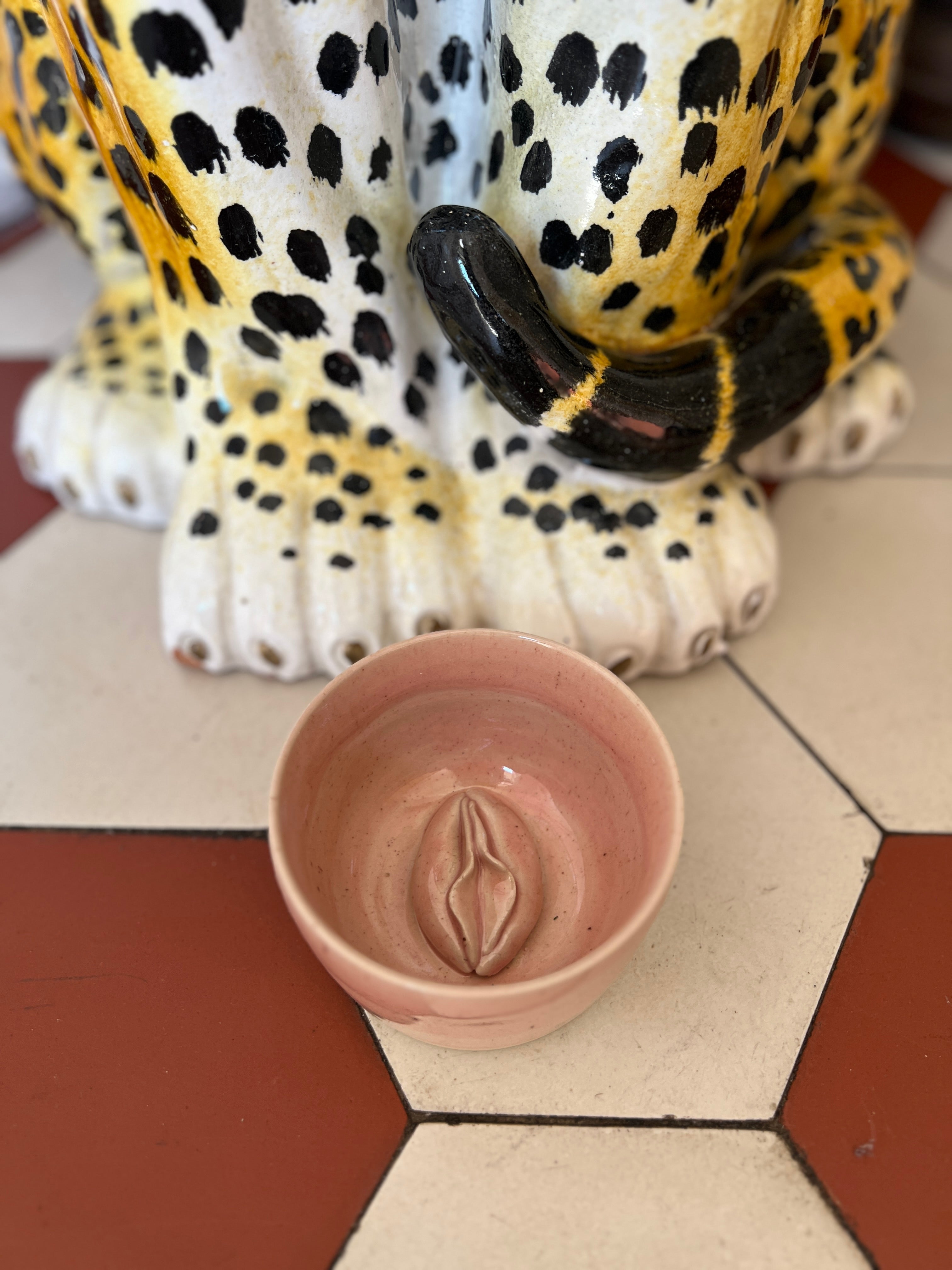 Ceramic bowl with a vulva design in front of a ceramic leopard statue on a tiled floor.